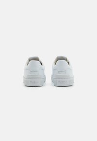 Zapatillas de piel blanca con una textura suave, forma redondeada y suela de goma. Presentan un logo en el talón y elementos de diseño minimalistas.
