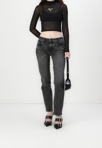 Vrouw in een zwarte doorschijnende cropped blouse met lange mouwen en tekst, vervaagde zwarte spijkerbroek, puntige zwarte gespen schoenen, met een kleine zwarte handtas.