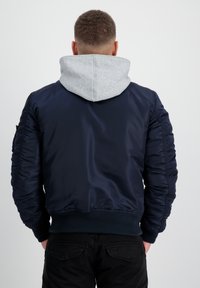 Jachetă de bomber bleumarin cu glugă gri, fabricată dintr-un material neted. Are manșete și tiv cu aspect canalat, precum și mâneci pufoase pentru un design structurat.