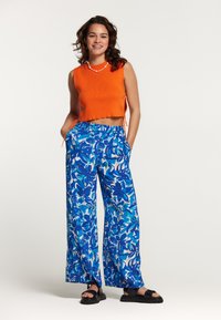 Narančasti crop top s rebrastom teksturom i bočnim vezicama; plave hlače širokih nogavica s cvjetnim uzorkom; crne sandale.