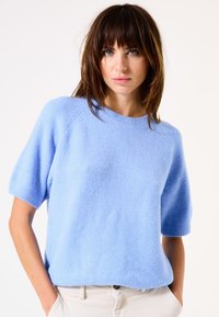 Helles blaues, kurzärmliges Sweatshirt aus weichem, strukturiertem Material mit einem gerippten Halsausschnitt und gesticktem Saum, kombiniert mit hellen Hosen.