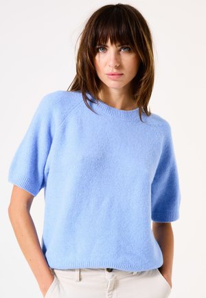 Pull bleu clair à manches courtes en matière douce et texturée avec un col côtelé et un ourlet cousu, associé à un pantalon de couleur claire.