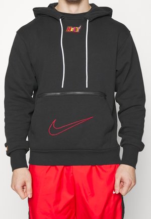 Uomo che indossa una felpa nera Nike con logo "Heat" rosso, cordini bianchi, tasca con cerniera frontale e pantaloncini Nike rossi.