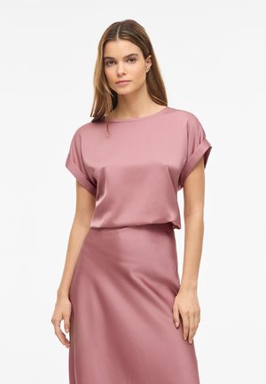 Mujer con cabello largo que lleva una blusa de satén rosa empolvado de manga corta y falda a juego, de pie frente a un fondo blanco liso.