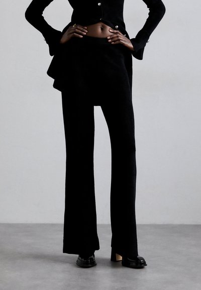 Μαύρο κομμένο κοστούμι με εφαρμοστό κοντό σακάκι με ασημί κουμπιά και παντελόνι με φαρδιές πλύσεις. Το ύφασμα έχει λεία υφή με διακριτική γυαλάδα.