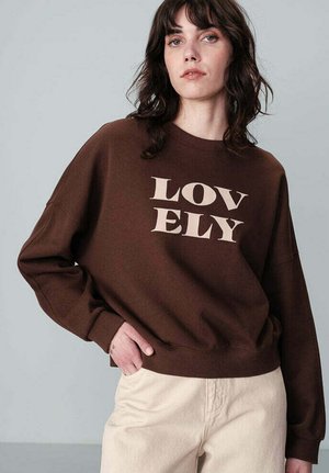 Sweatshirt marron avec le texte « LOVELY » en beige clair, encolure ronde, coupe décontractée et coutures d'épaule tombantes, associé à un pantalon couleur crème.