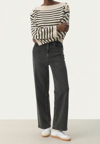 Raidallinen neulepaita kermanvalkoisena ja mustana, leveälahkeiset tummat farkut ja valkoiset lenkkarit harmailla yksityiskohdilla, kaikki kuvattu teksturoidulla pinnalla.