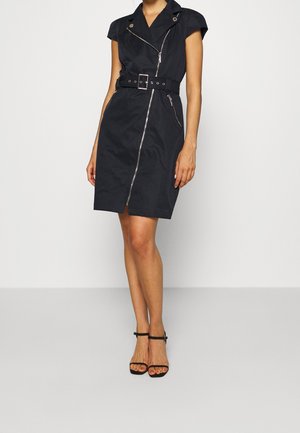 Frau trägt ein ärmelloses schwarzes Kleid mit Gürtel und diagonalen Reißverschlussdetails vorne sowie schwarze, hochhackige Sandalen mit Knöchelriemen.