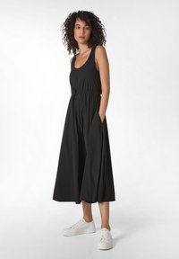 Abito nero senza maniche con vestibilità ampia, vita cin cin, e tasche laterali. Abbinato a sneakers bianche, che mettono in risalto un design casual e fluido.