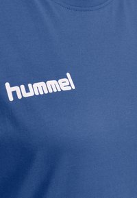 Camiseta de algodón azul con el logotipo blanco "hummel" en el frente, con un cuello redondo y una textura de tela suave.