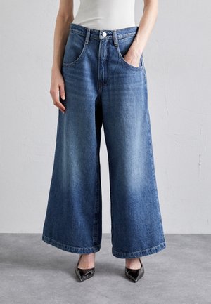 Kvinde iført blå denim jeans med brede ben, hvid ærmeløs top og sorte spidse hæle, stående mod en ensfarvet væg.