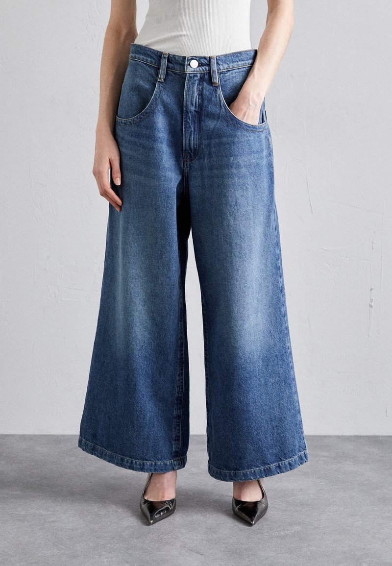 Femme portant un jean denim bleu à jambes larges, un haut blanc sans manches et des talons noirs pointus, debout contre un mur uni.