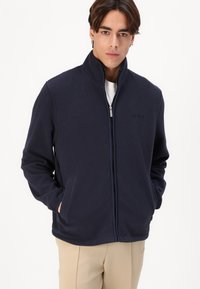 Jachetă fleece de culoare bleumarin, cu guler înalt, fermoar frontal și buzunare laterale. Prezintă un material texturat și un mic logo pe piept.