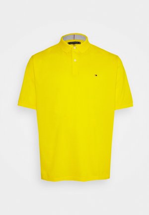 Polo jaune vif à manches courtes avec patte de boutonnage à deux boutons et petit logo drapeau sur la poitrine gauche sur fond blanc.