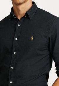 Schwarze Baumwollbluse mit strukturiertem Kragen, ausgestattet mit einem kleinen goldenen Polo-Logo auf der linken Brust und weißen Knöpfen.