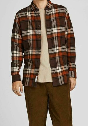 Homme portant une chemise à carreaux marron, orange et blanche ouverte sur un t-shirt beige, avec un pantalon en velours côtelé marron et des bagues en argent aux doigts.