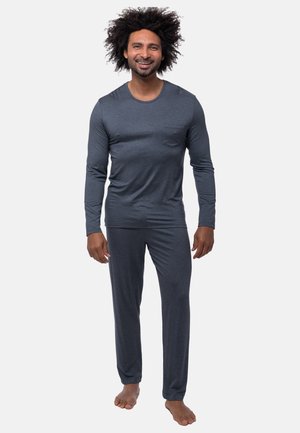 Uomo con capelli ricci in piedi scalzo, indossa una maglia a maniche lunghe grigio scuro e pantaloni abbinati su uno sfondo semplice.