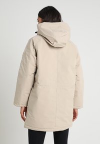 Abrigo acolchado beige con capucha, de corte recto, mangas largas y una textura suave y mate. Sin patrones visibles ni detalles de hardware.