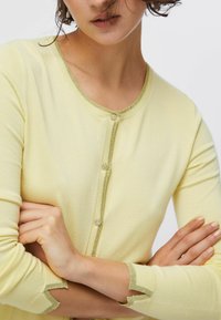 Nő világos sárga kardigánban, arany szegéllyel és díszített gombokkal, karjai keresztbe téve, haja részben az arcát takarja, fehér háttér előtt.