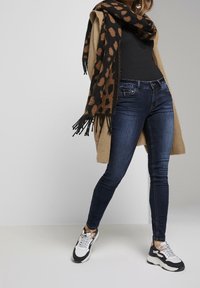 Jeans ajustados de color azul oscuro combinados con una camiseta negra de canalé, un cárdigan beige y una bufanda de estampado de leopardo. Zapatillas deportivas blancas y negras completan el conjunto.