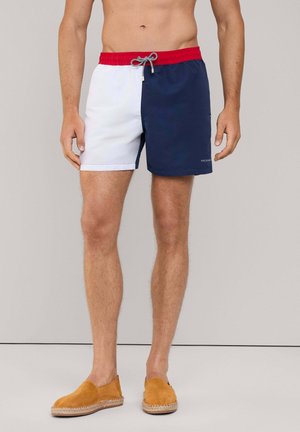 Pantaloncini da bagno bianchi e blu navy, con una vita elastica rossa con cordoncino, realizzati in tessuto morbido, lunghi fino al ginocchio e dotati di tasche laterali.