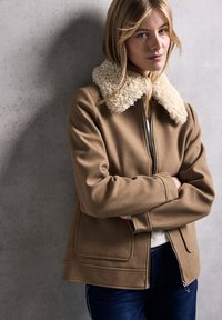 Giacca in pile marrone con colletto in pelliccia sintetica color crema, chiusura con zip, tasche laterali e una vestibilità leggermente ampia, abbinata a jeans scuri.