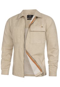 Chemise beige à manches longues avec deux poches avant, fermeture éclair sur le devant et un accent orange le long de la fermeture. Fabriquée en tissu texturé.