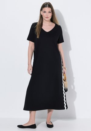 Kvinde iført en sort midi-kjole med korte ærmer og hvide bølgede striber på siderne, der holder en flettet håndtaske med træhåndtag.