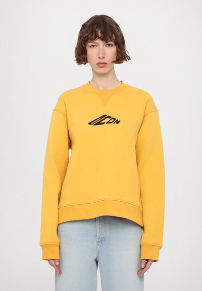 Gul sweatshirts med rund halsudskæring, der har et sort logo på brystet. Lange ærmer, afslappet pasform og synlige syninger.