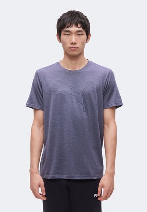 Ung mand iført en grå Puma T-shirt med et diskret sort Puma-logo og sorte Puma-bukser, stående foran en ensfarvet baggrund.