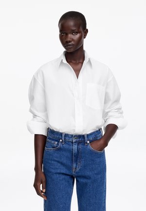 Vrouw draagt een loszittend wit overhemd met opgerolde mouwen, ingestopt in een high-waisted blauwe spijkerbroek, hand in de zak, tegen een witte achtergrond.