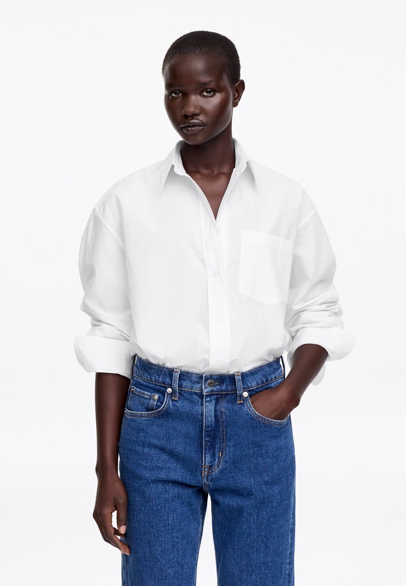 Femme portant une chemise blanche ample à boutons avec les manches retroussées, rentrée dans un jean bleu taille haute, main dans la poche, sur fond blanc.