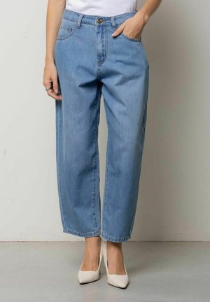 Persona che indossa jeans azzurri a gamba dritta, tacchi bianchi alti, con la mano destra in tasca e la mano sinistra rilassata al fianco.