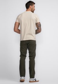 Beige t-shirt met korte mouwen, gecombineerd met olijfgroene broek en witte sneakers met zwarte accenten. Tatoeages zichtbaar op de onderarm.