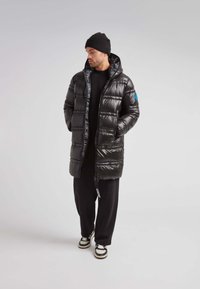 Hombre de pie con las manos en los bolsillos, vestido con un abrigo acolchado negro largo, un gorro negro, pantalones negros y zapatillas en blanco y negro.