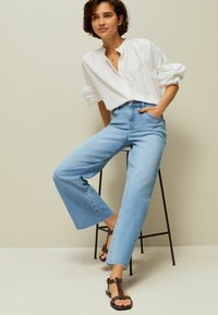 Jeans de cintura alta de color azul claro con pierna acampanada, combinados con una camisa blanca de botones con mangas abullonadas y sandalias marrones.