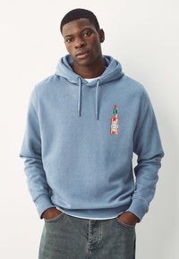 Mand iført lyseblå hættetrøje med et lille Tabasco-flaske grafik på brystet, hænderne i lommerne, stående foran en hvid baggrund.