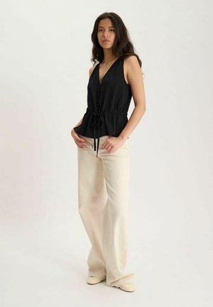 Femme debout portant un haut noir sans manches boutonné avec taille à cordon et un pantalon beige à jambes larges sur un fond uni.