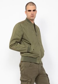 Chaqueta bomber verde oliva con puños y dobladillo acanalados, que presenta un cierre de cremallera y bolsillos laterales. Llevada con pantalones cargo a juego.