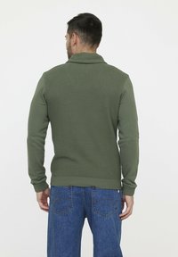 Grüner strukturierter Pullover mit geripptem Saum und Bündchen, kombiniert mit blauen Jeans. Die Rückseite hat einen Kragen und dezente Details auf dem Stoff.