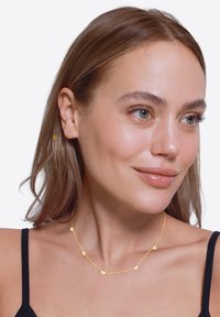 Collier en or délicat avec des accents en forme de cœur texturés, doté d'une fine chaîne et d'un design lisse et minimaliste. Porté sur un haut sombre.
