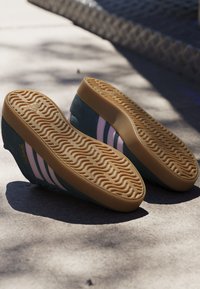 Paire de baskets Adidas avec des rayures roses et des semelles en gomme naturelle, faces vers le haut sur une surface en béton avec des motifs d'ombres.