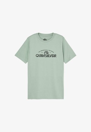 Chłopiec o kręconych włosach, w jasnooliwkowej koszulce Quiksilver, czarnych spodniach i czarnych trampkach, stojący na jednolitym tle.