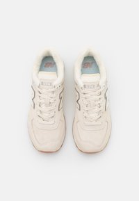 Pár béžových tenisek New Balance 574 s bílým fleecem uvnitř, šňůrky zavázané, pohled shora na bílém pozadí.