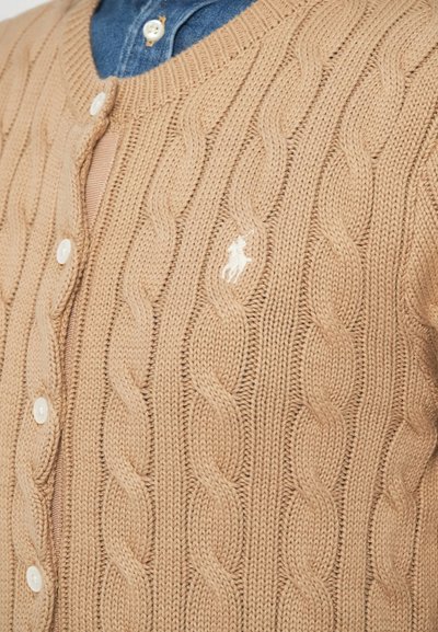 Adīts kardigans gaiši bēšā krāsā ar pinuma rakstu, kam ir V veida kakla izgriezums, pogu aizdare un neliels izšūts logotips uz krūtīm.