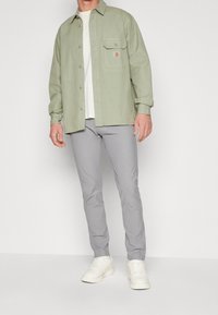 Camicia verde chiaro con bottoni e taschino sul petto, abbinata a pantaloni grigi slim-fit e sneakers bianche. Tessuto liscio, design casual, stile minimalista.