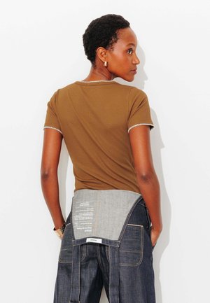 Femme portant un haut marron à manches courtes et un pantalon en denim foncé avec un détail en denim clair, debout les mains dans les poches, de profil sur un fond blanc.