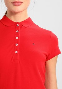 Röd polo tröja gjord av strukturerad tyg, med klassisk krage, korta ärmar och en logotypaccent på vänster bröst med knappknäppning.