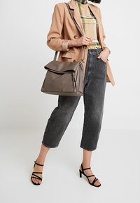 Borsa in pelle grigia con patta pieghevole e dettagli con cerniera. Il modello indossa un blazer beige, una camicia a quadri, jeans neri a vita alta e scarpe con tacco nere.