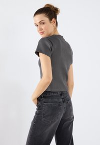 Grå ribbet kortermet topp kombinert med høyt liv svarte jeans. Modellen står med hendene i lommene, vendt litt bakover med en nøytral bakgrunn.